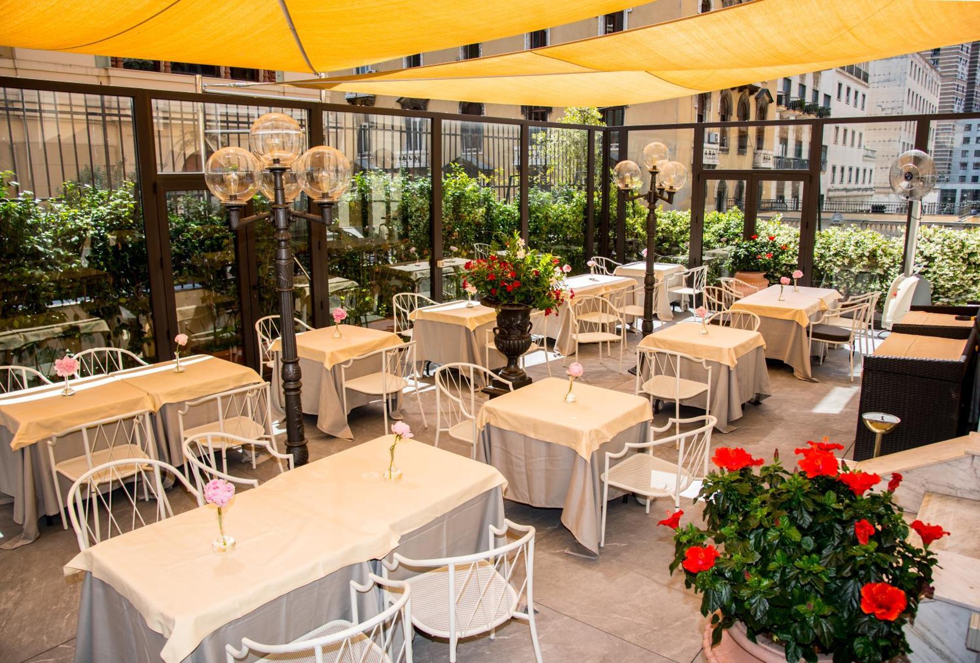 Hotel Bristol Palace Genoa Exterior photo The photo shows an outdoor dining area, featuring several tables covered with light-colored tablecloths. Each table has a flower vase placed on it, adding a touch of decoration. The seating consists of white chairs, and the area is enclosed with glas