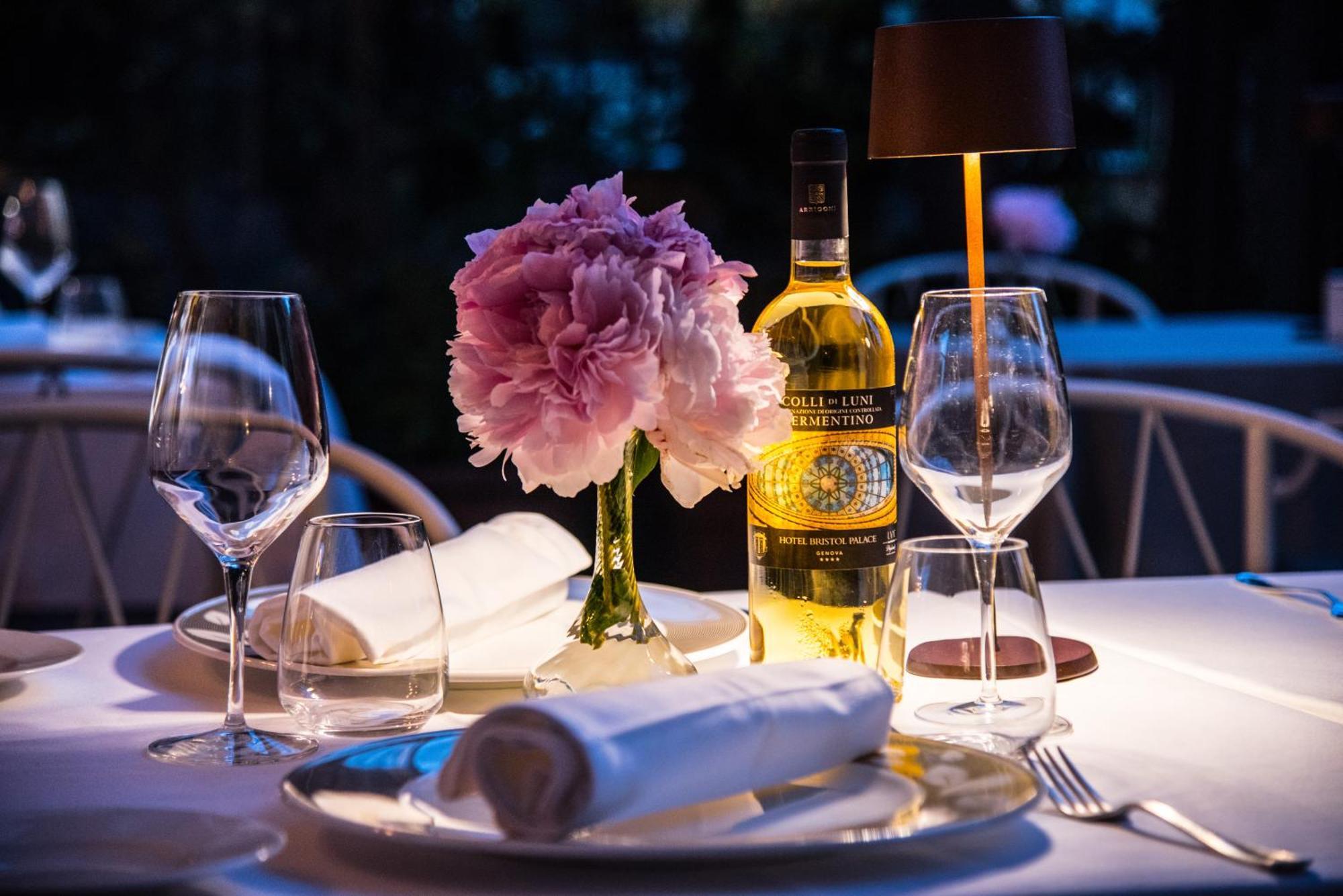 Hotel Bristol Palace Genoa Exterior photo The photo shows a beautifully set dining table with an elegant ambiance. There is a vase with a pink flower arrangement at the center, adding a touch of color and freshness. A bottle of white wine is also prominently displayed next to the flowers. Th