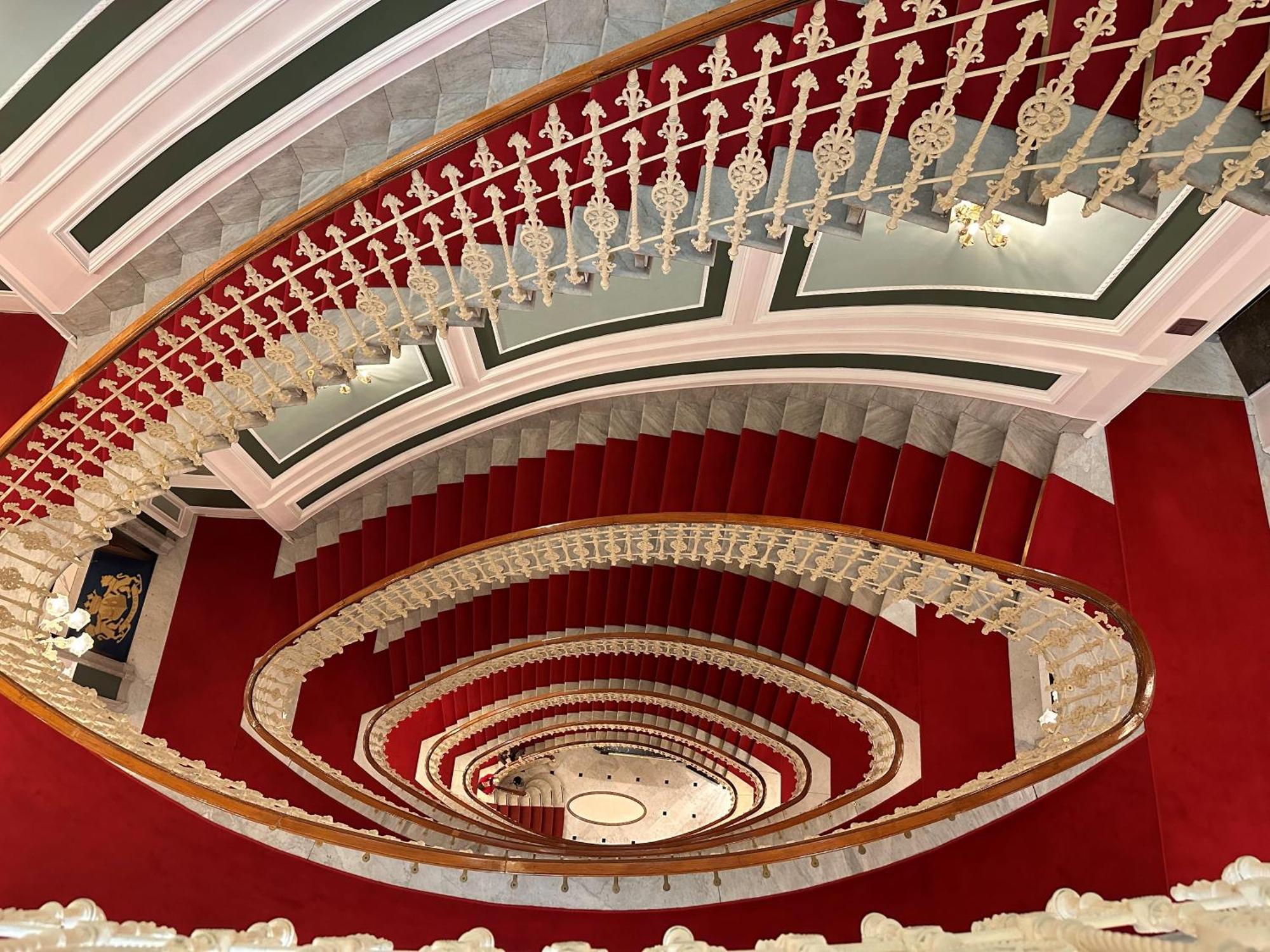 Hotel Bristol Palace Genoa Exterior photo The photo shows a grand, spiral staircase viewed from above. The staircase features elegant white railings with intricate designs and is surrounded by a rich red carpet. The walls have a classic architectural style, with decorative elements in soft c