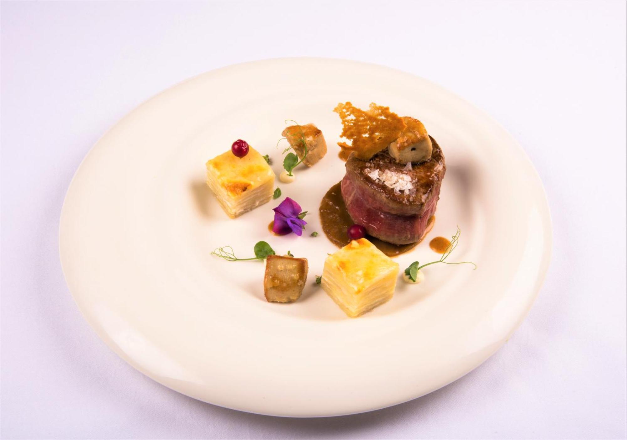 Hotel Bristol Palace Genoa Exterior photo The photo shows an elegantly plated dish featuring a central piece of meat wrapped in a savory outer layer, possibly prosciutto or bacon, topped with a sauce or garnishment. Surrounding the main protein are small cubes that appear to be a starch, lik