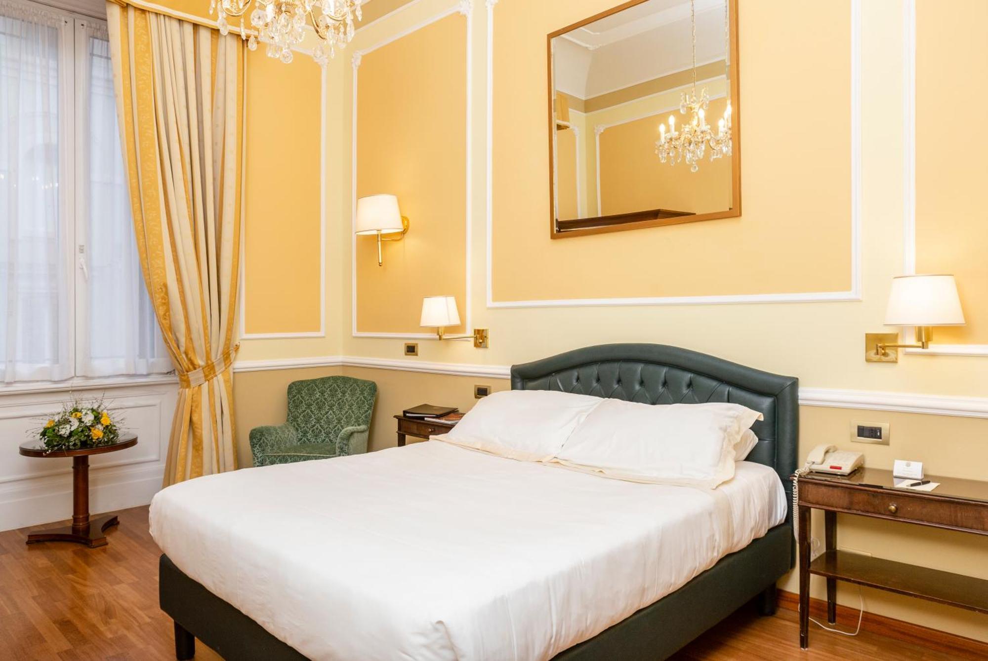Hotel Bristol Palace Genoa Room photo The photo shows a cozy hotel room with a warm color scheme. It features a large bed with a white duvet, neatly arranged. To the left, there's a light green armchair and a small table. The walls are painted a soft yellow, and there are elegant curtain