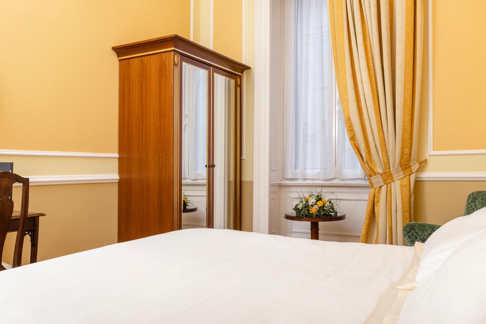 Hotel Bristol Palace Genoa Room photo The photo shows a cozy bedroom with warm yellow walls. There is a bed with white bedding in the foreground. On one side of the room, there is a wooden wardrobe that features a mirrored door. Beside it, there's a small table adorned with a floral arra