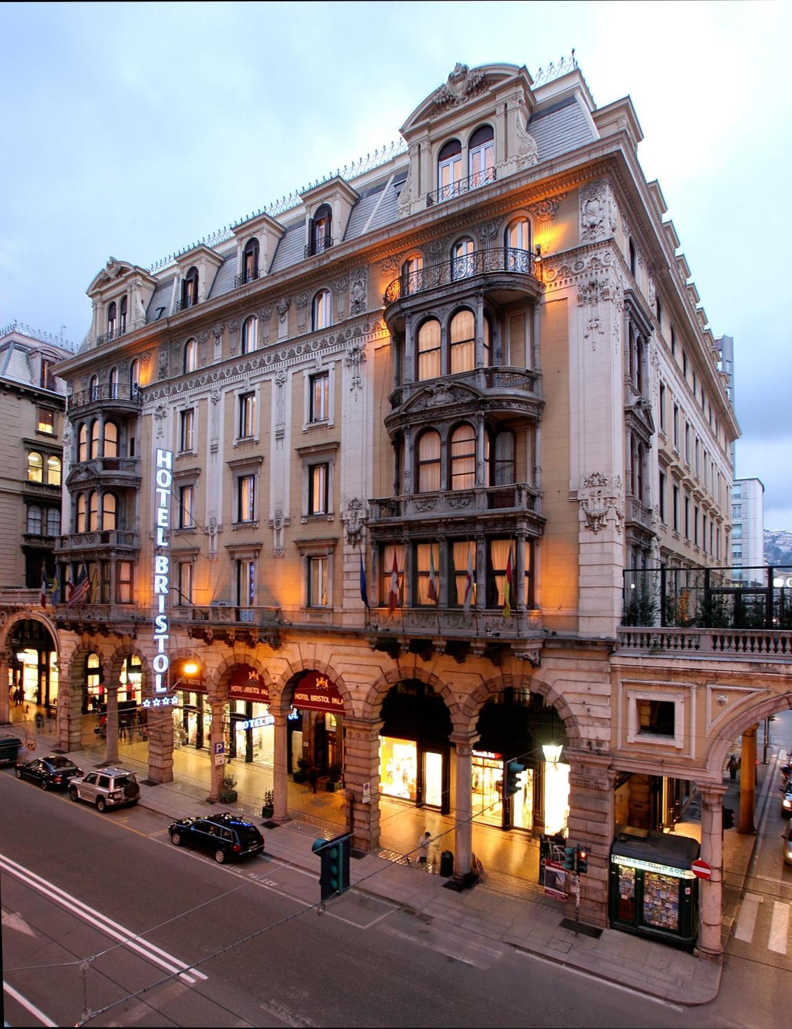 Hotel Bristol Palace Genoa Exterior photo The photo shows a grand hotel building, likely the Hotel Bristol. It features an elegant architectural design with ornate details, including balconies and decorative moldings. The façade is illuminated, giving it a warm glow in the evening light. The
