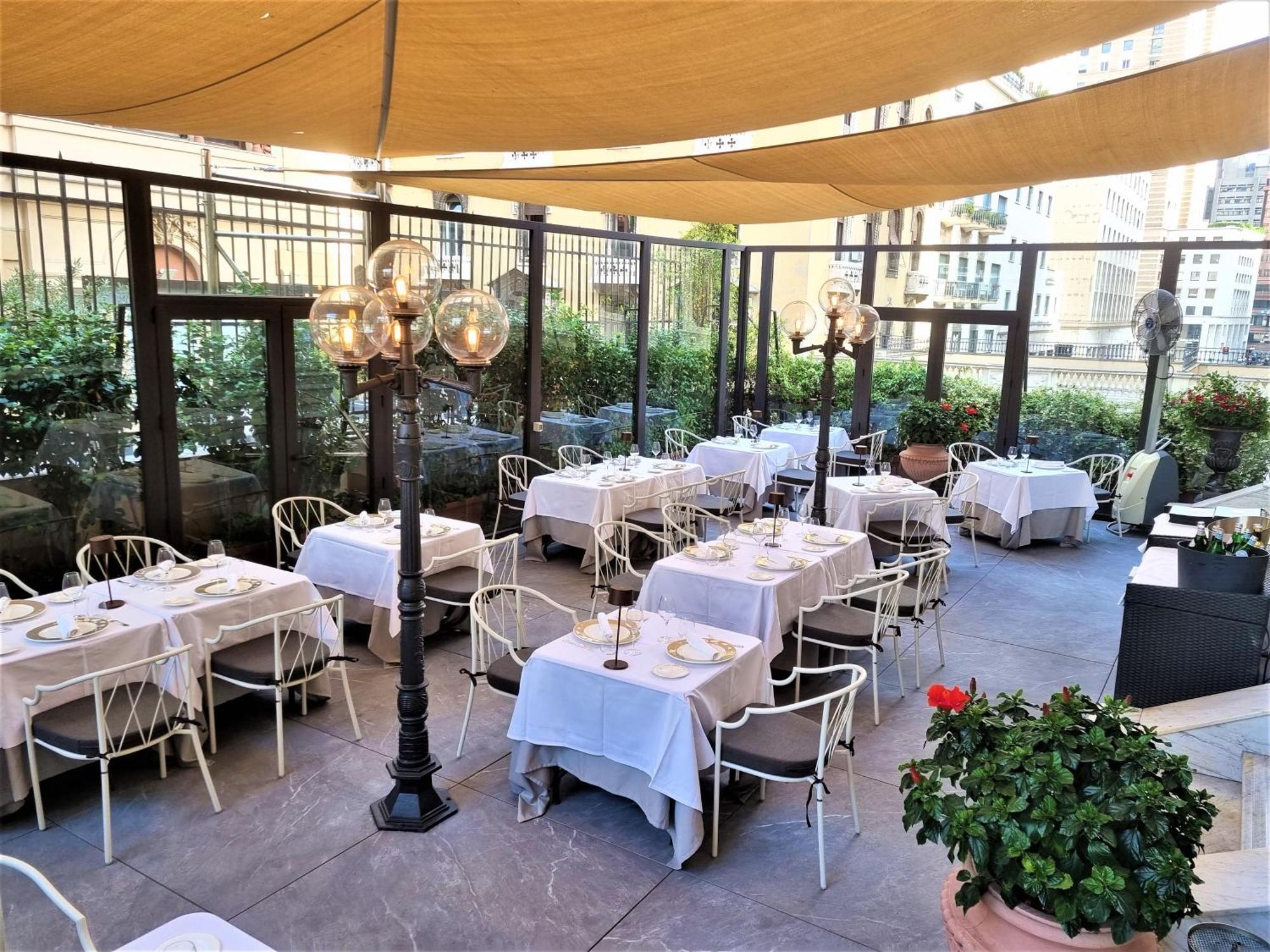 Hotel Bristol Palace Genoa Exterior photo The photo shows an outdoor dining area of a restaurant. There are several neatly arranged tables covered with white tablecloths, set with plates and utensils. The space is adorned with decorative lanterns on tall stands, providing a cozy atmosphere. 