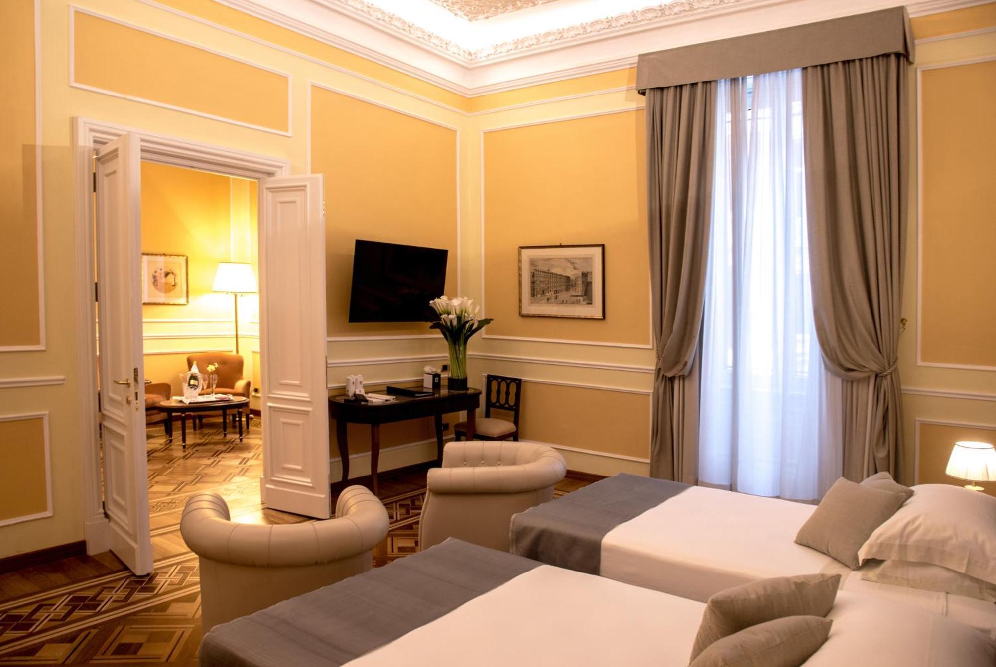 Hotel Bristol Palace Genoa Exterior photo The photo shows an elegantly designed hotel room with soft yellow walls and high ceilings. There are two beds with gray covers positioned in the foreground. The room features large windows with flowing curtains that allow natural light to enter. In t