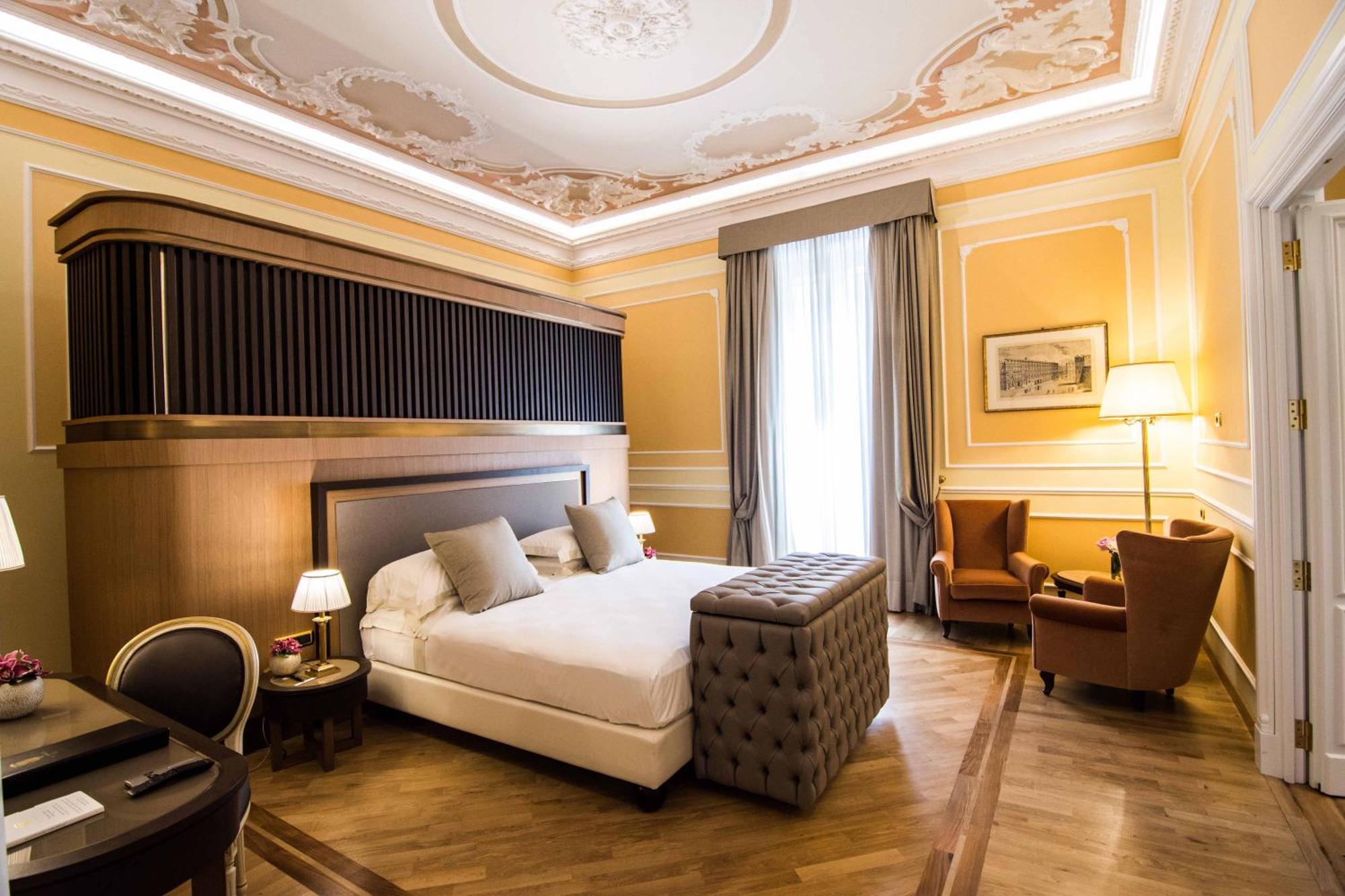 Hotel Bristol Palace Genoa Exterior photo The photo shows an elegantly designed hotel room. It features a large bed with plush pillows, and a tufted ottoman at the foot of the bed. The walls are painted in a warm yellow tone, adorned with decorative molding. A pair of tall windows are draped