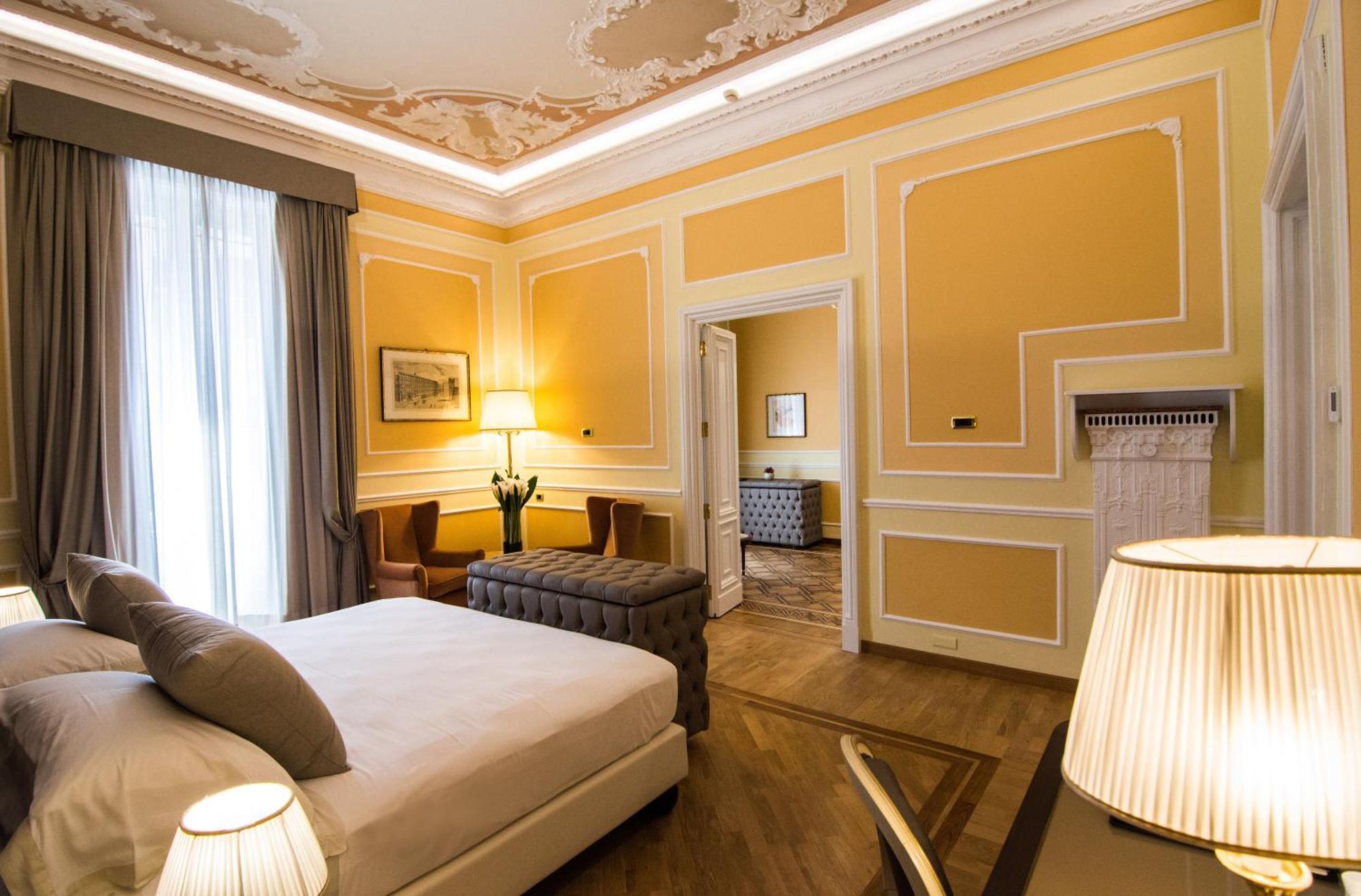 Hotel Bristol Palace Genoa Exterior photo The photo shows a well-decorated hotel room with a warm yellow color scheme. There is a large bed with neutral-toned bedding and pillows. To the side, there are two armchairs and a small table, creating a cozy seating area. A stylish lamp sits on a n