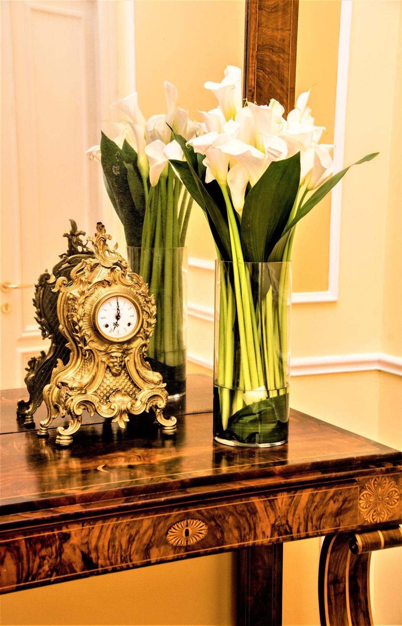 Hotel Bristol Palace Genoa Exterior photo The photo features an ornate vintage clock with a decorative gold finish, placed on a wooden surface. Next to the clock is a tall, clear vase filled with elegant white calla lilies and green leaves. The background shows a softly lit interior with lig