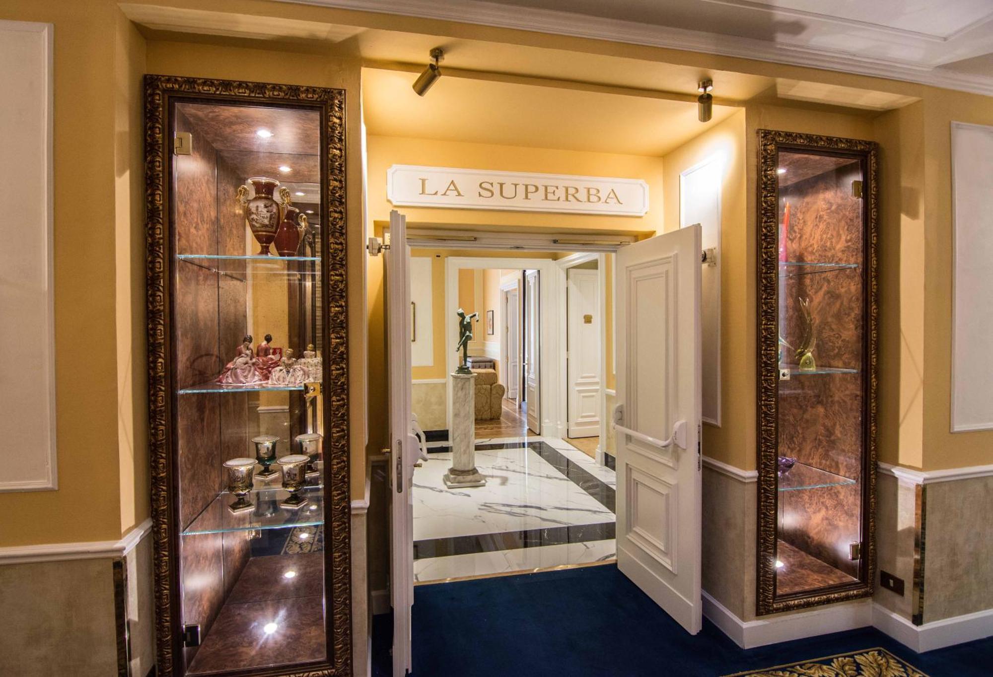 Hotel Bristol Palace Genoa Exterior photo The photo shows the entrance to a venue or restaurant named "La Superbia." The entrance features double doors that are open, revealing an elegant interior. Flanking the entrance are glass display cases with ornate decorations, which likely showcase f