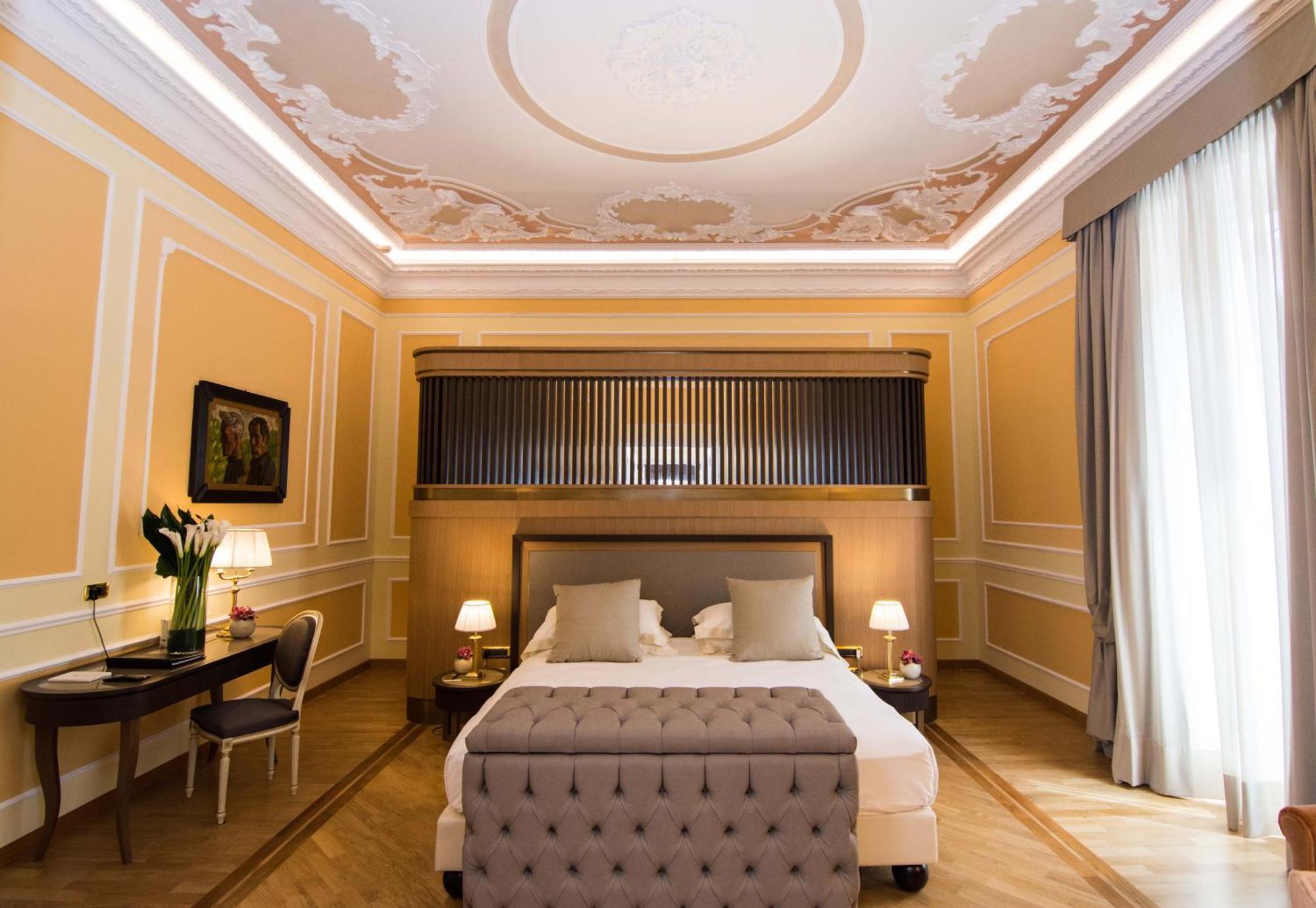 Hotel Bristol Palace Genoa Exterior photo The photo shows a stylish and elegant bedroom. The room features light peach-colored walls with ornate moldings. The ceiling has decorative elements, creating an upscale atmosphere. In the center, there is a large, gray upholstered bed with plush bed