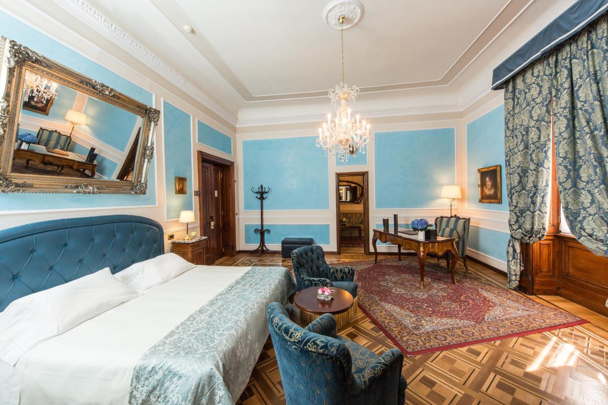 Hotel Bristol Palace Genoa Room photo The photo shows a beautifully designed hotel room or suite. It features light blue walls with decorative molding, providing an elegant ambiance. The room includes a large bed with a gray coverlet, accompanied by plush blue armchairs and a round table