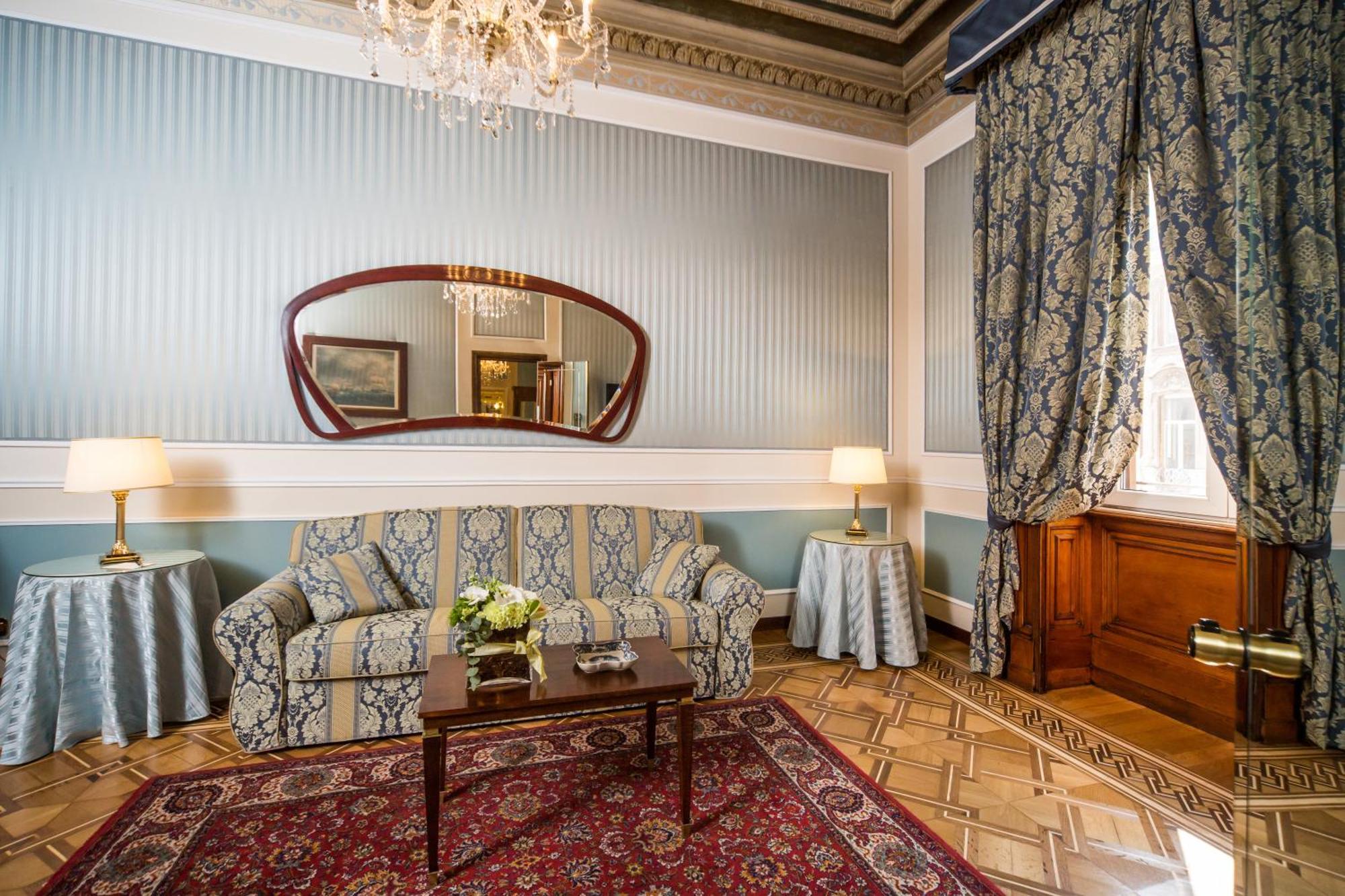 Hotel Bristol Palace Genoa Room photo The photo shows an elegant living room featuring classic decor. The walls are painted in a soft blue with vertical stripes, and ornate crown molding adds to the room's charm. A large mirror with a wavy frame hangs above a patterned couch that complem