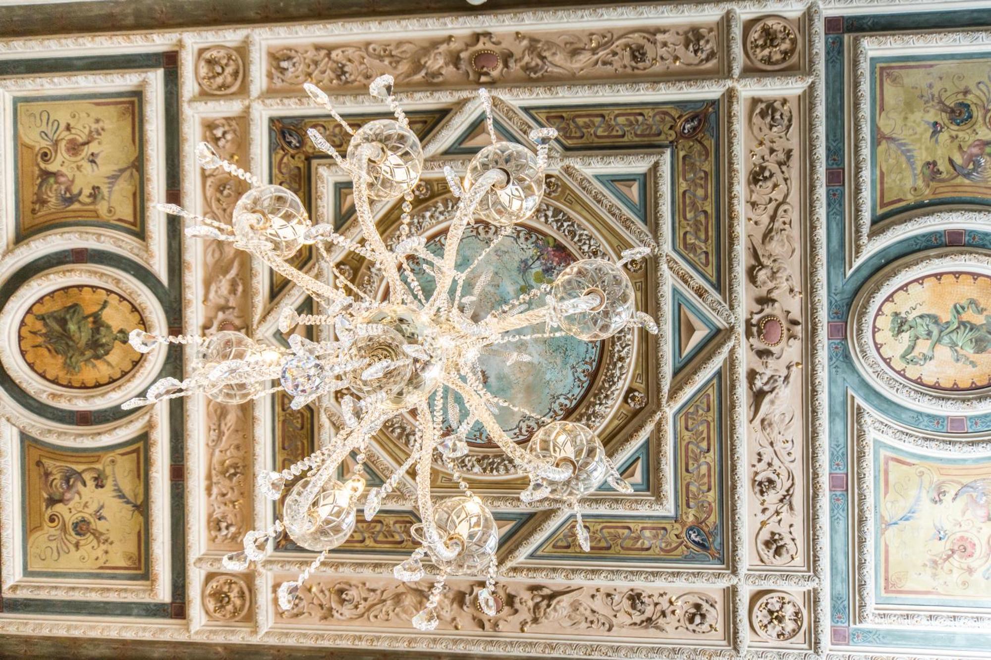 Hotel Bristol Palace Genoa Room photo The image depicts an ornate ceiling featuring a large chandelier made of crystal. The chandelier hangs from a richly detailed ceiling adorned with intricate designs, including decorative molding and colorful frescoes. The artwork includes circular mo